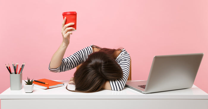 Woman with cup raised, head on table next to computer
